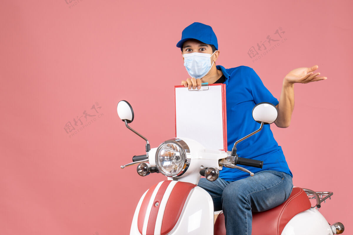 桃顶视图好奇的男性送货员戴着面具戴着帽子坐在滑板车上 展示粉彩桃色背景下的文件显示滑板车男
