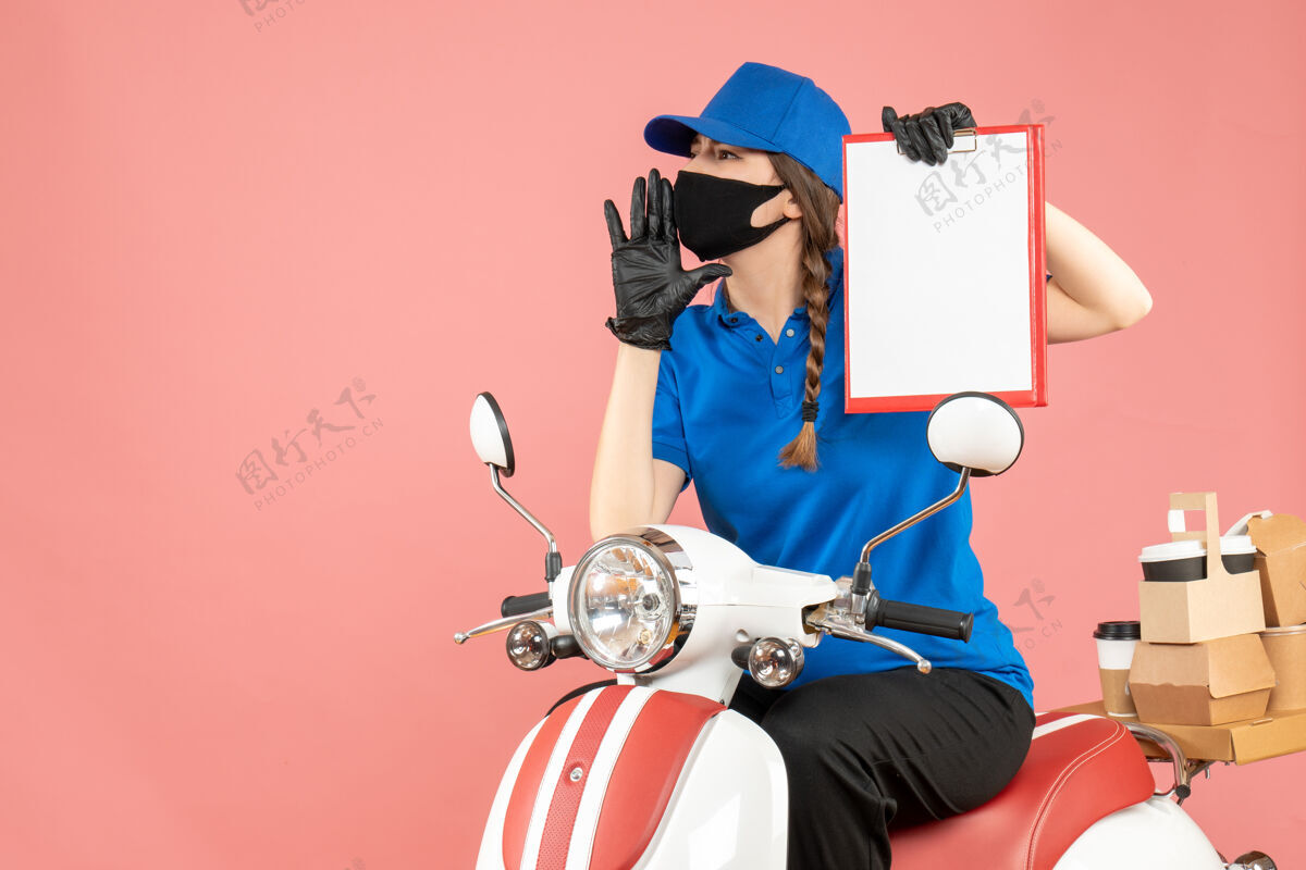 坐俯视图紧张的快递女孩戴着医用口罩和手套坐在滑板车上拿着空纸在粉彩桃色背景下传递订单桃纸举行