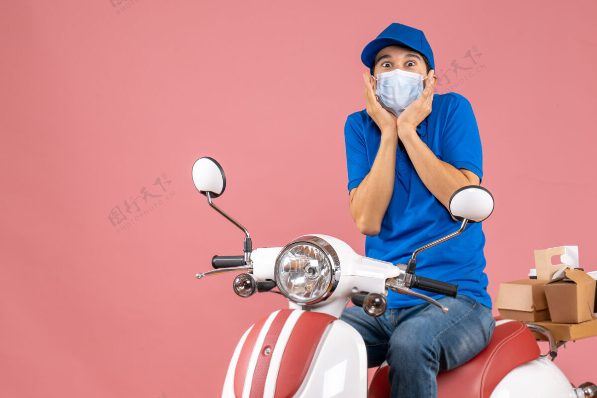 医疗戴着医用面罩的送货员戴着帽子坐在滑板车上 在柔和的桃色背景下感到惊讶摩托车桃面具
