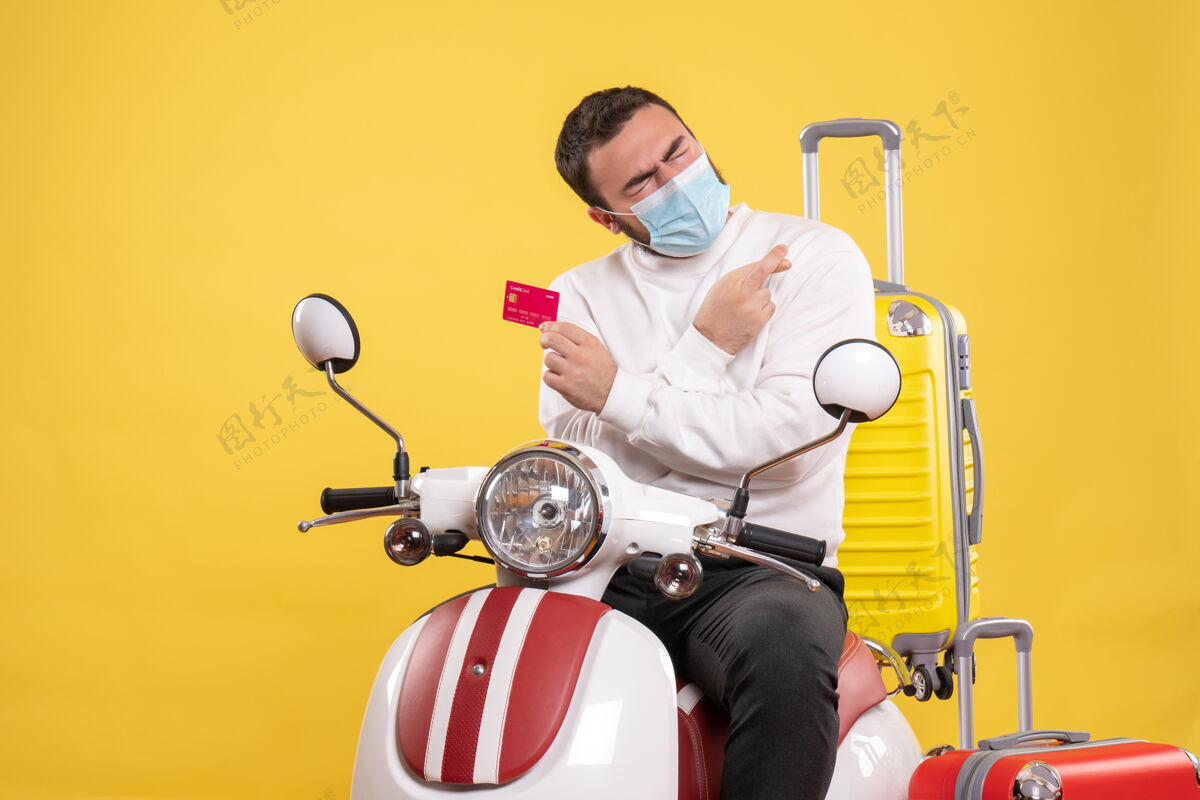 抱着旅行概念的正面图 戴着医用面罩的年轻人坐在摩托车上 黄色手提箱 手里拿着银行卡面罩车辆黄色