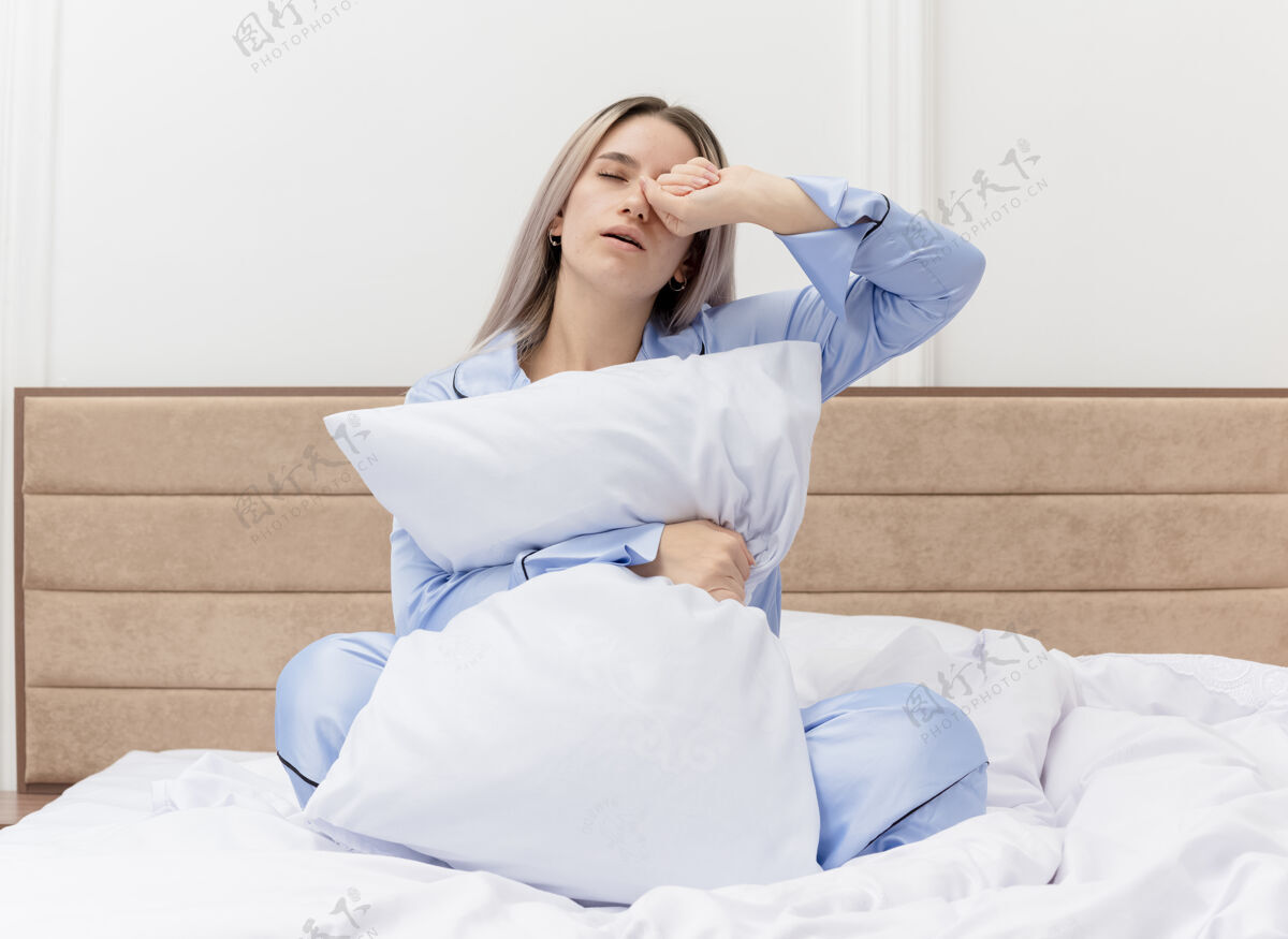 枕头穿着蓝色睡衣的年轻美女 带着枕头坐在床上醒来 卧室里感到晨曦疲惫睡衣灯光卧室