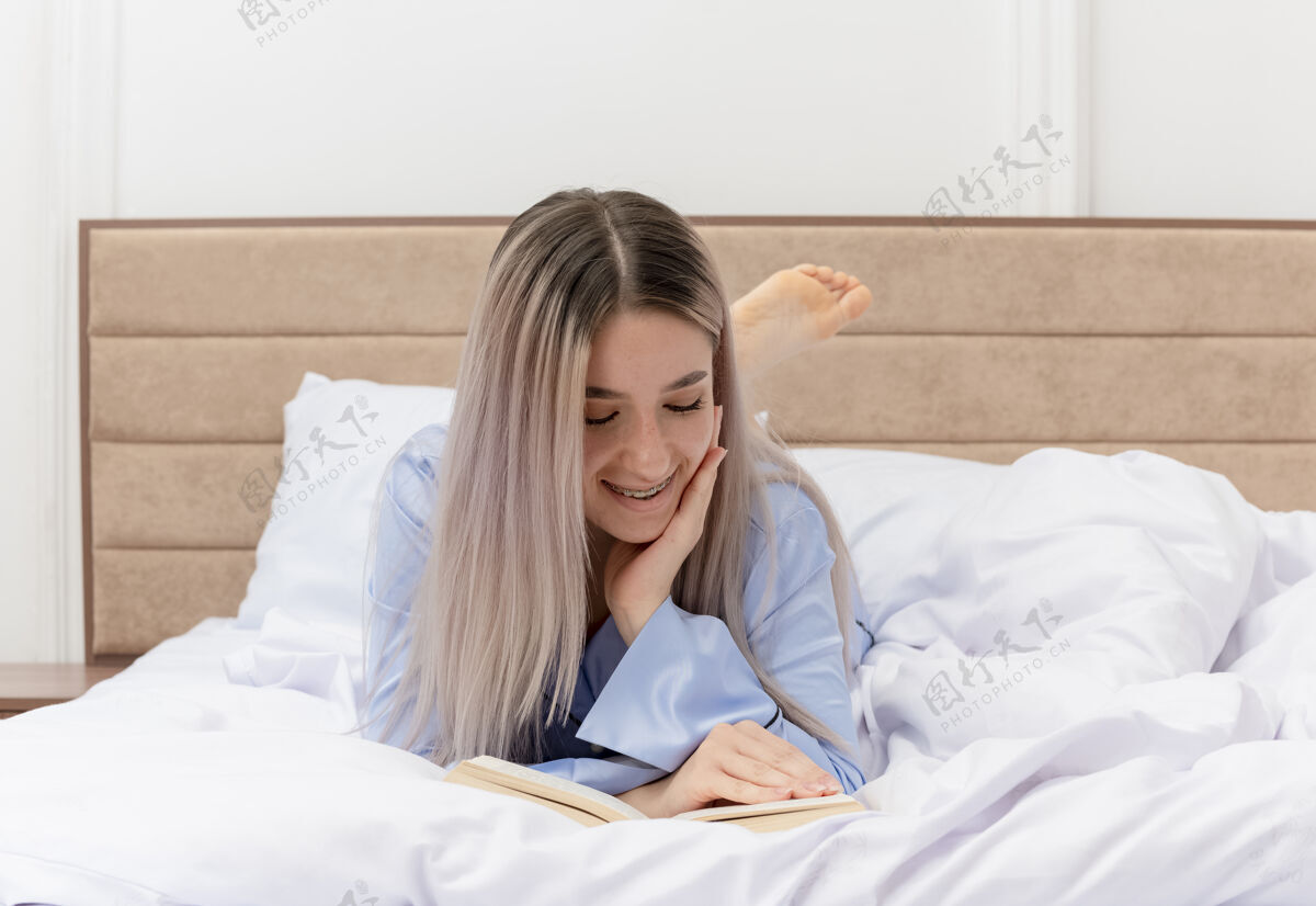 睡衣穿着蓝色睡衣的年轻美女躺在床上休息 在卧室里微笑着看书蓝色休息床