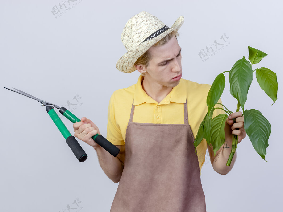 花园年轻的园丁穿着连体衣 戴着帽子 拿着树篱剪和植物 看着它很感兴趣剪子树篱站着