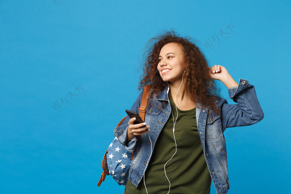 人年轻的非洲裔美国少女 穿着牛仔服 背着耳机 隔离在蓝色的墙上手机舞蹈青少年