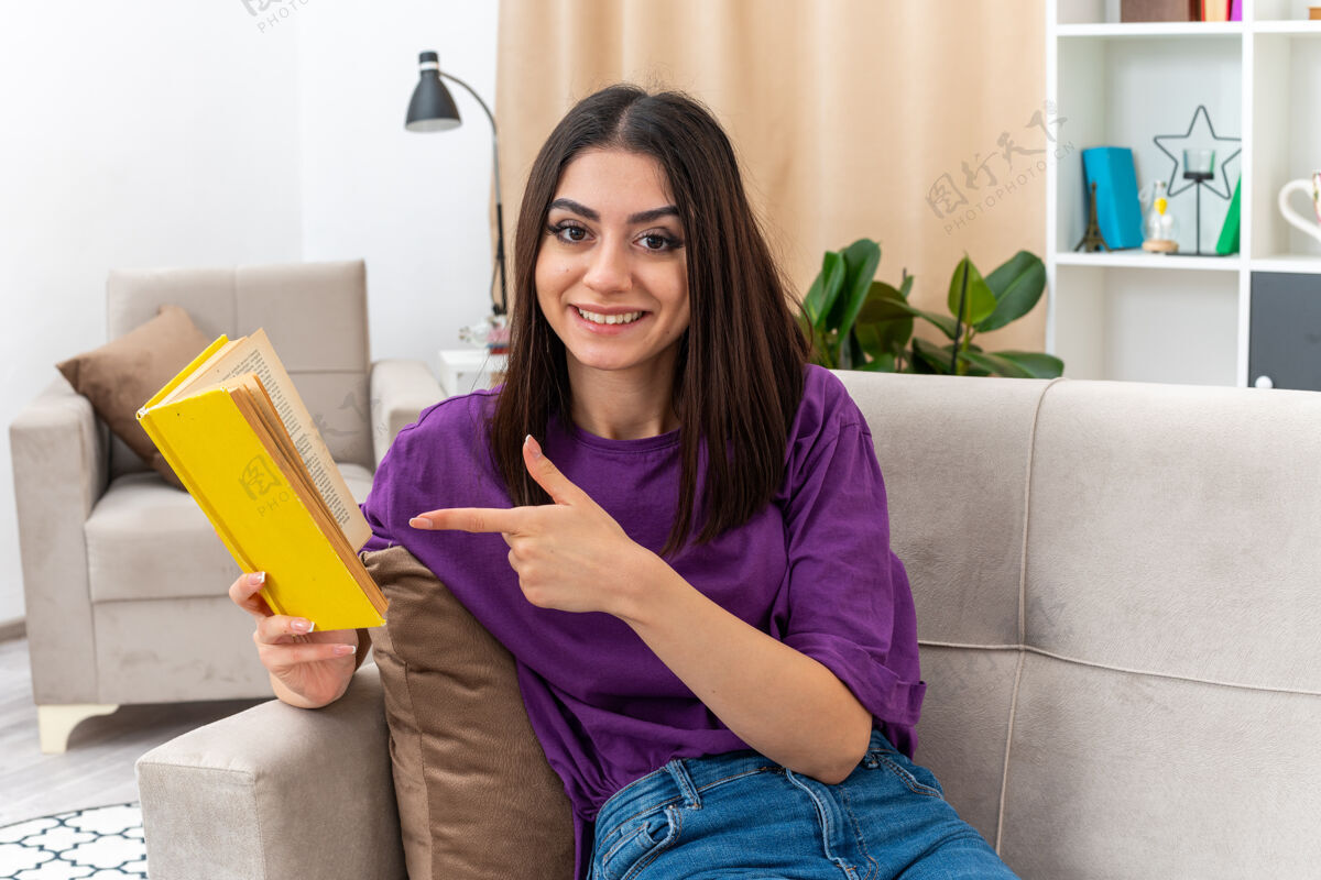 衣服穿着休闲服的年轻女孩拿着书 用食指指着书 快乐而积极地笑着 愉快地坐在客厅里的沙发上手指年轻休闲