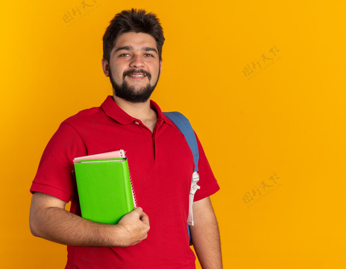 姿势年轻的留着胡子的学生 穿着红色马球衫 背着书包 面带微笑 站在橙色的墙上 快乐而自信胡须马球站立