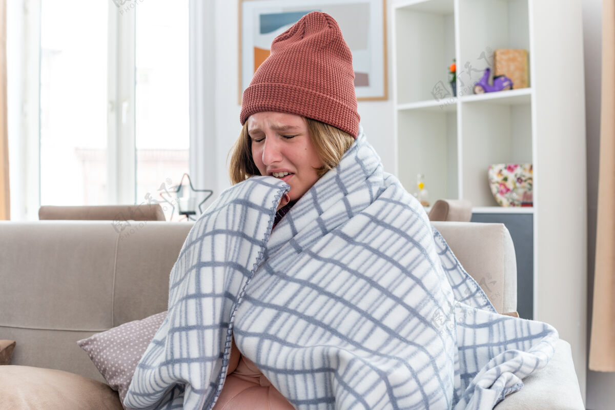 不适不健康的年轻女子 戴着暖和的帽子 裹着毯子 看起来不舒服 生病 感冒 流感 发烧 头痛 坐在客厅里的沙发上冷光痛苦