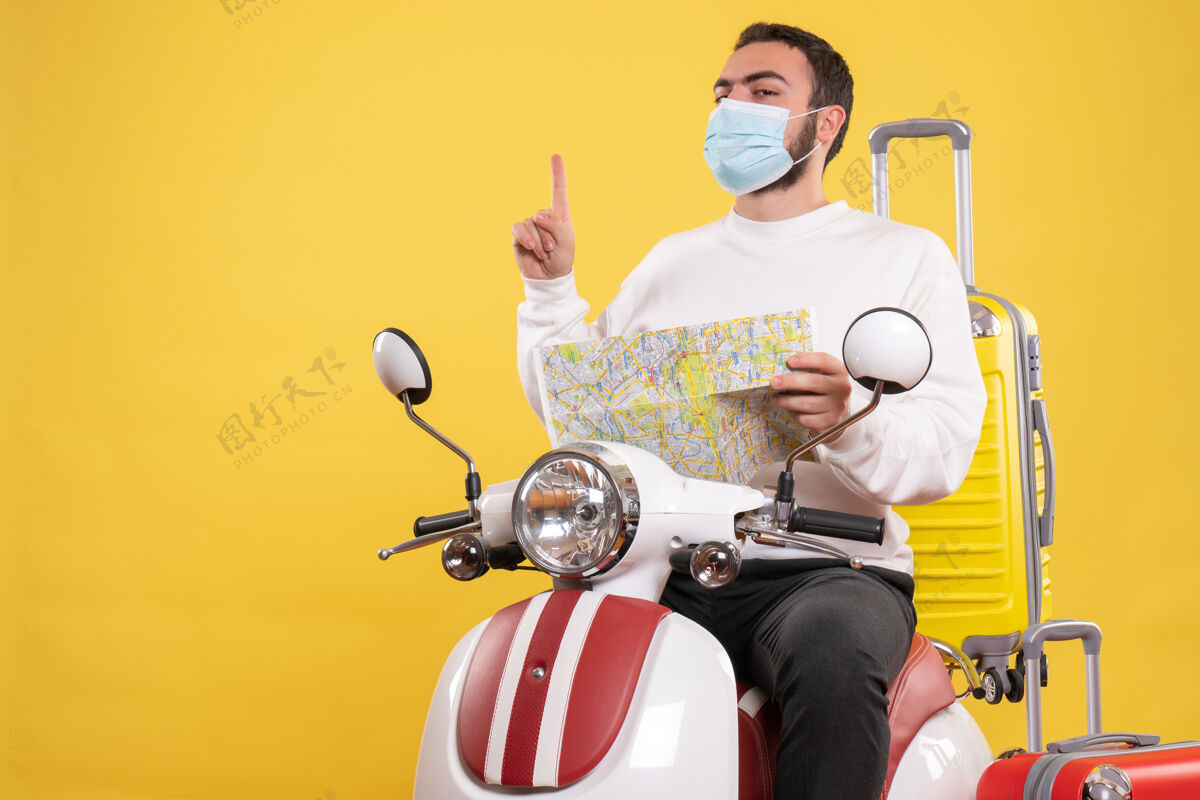 拿着旅行的概念与自信的家伙在医疗面具坐在摩托车上 黄色的手提箱上 并持有黄色的地图指向上坐着黄色旅行