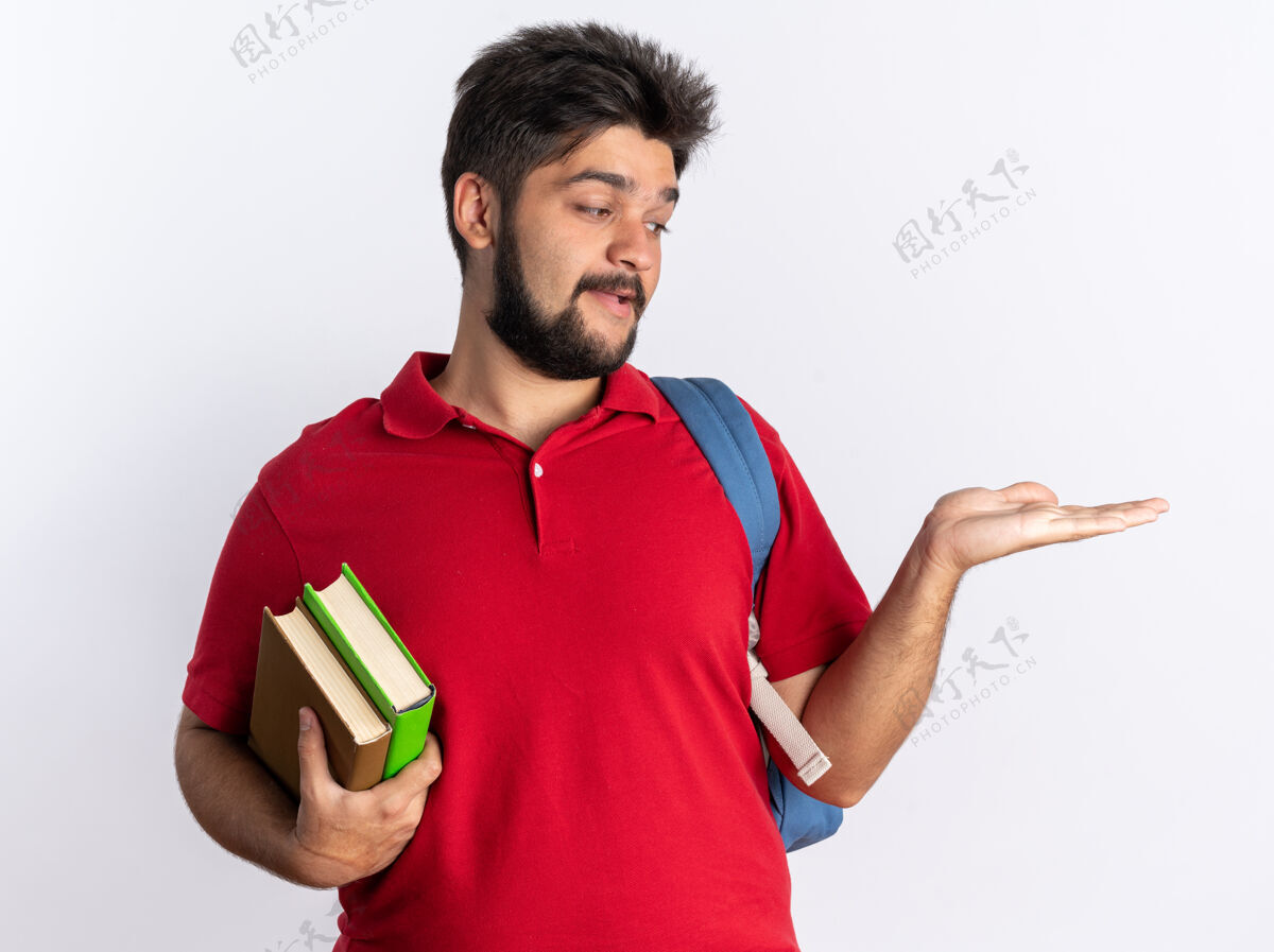 站立年轻的留着胡子的学生 穿着红色马球衫 背着书包 手拿笔记本 站在白墙上 快乐而积极地复制空间个人背包年轻人