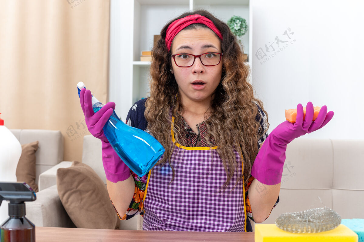 光戴着橡胶手套 拿着清洁喷剂和海绵的年轻女子坐在明亮的客厅里 手里拿着清洁用品和工具橡胶手套喷雾