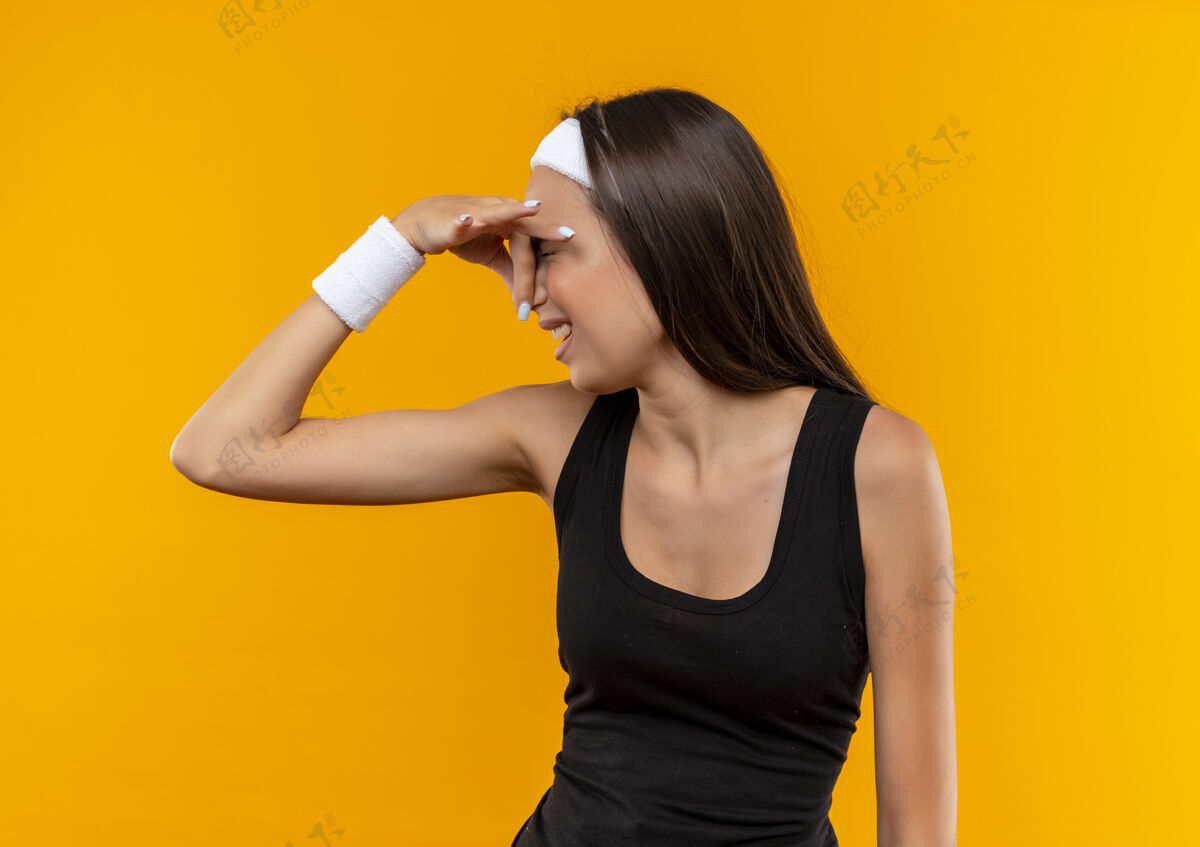 运动恼怒的年轻漂亮的运动女孩戴着头带和手环举行鼻子隔离在橙色墙上穿漂亮年轻