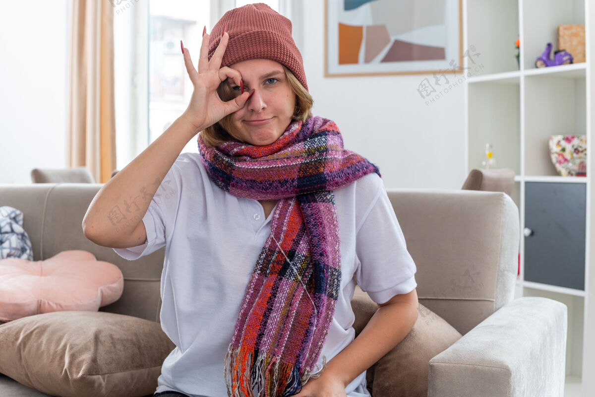 围巾不健康的年轻女子戴着帽子 脖子上围着暖和的围巾 患了感冒和流感 感觉好多了 做得很好 标志微笑着坐在客厅的沙发上温暖周围脖子