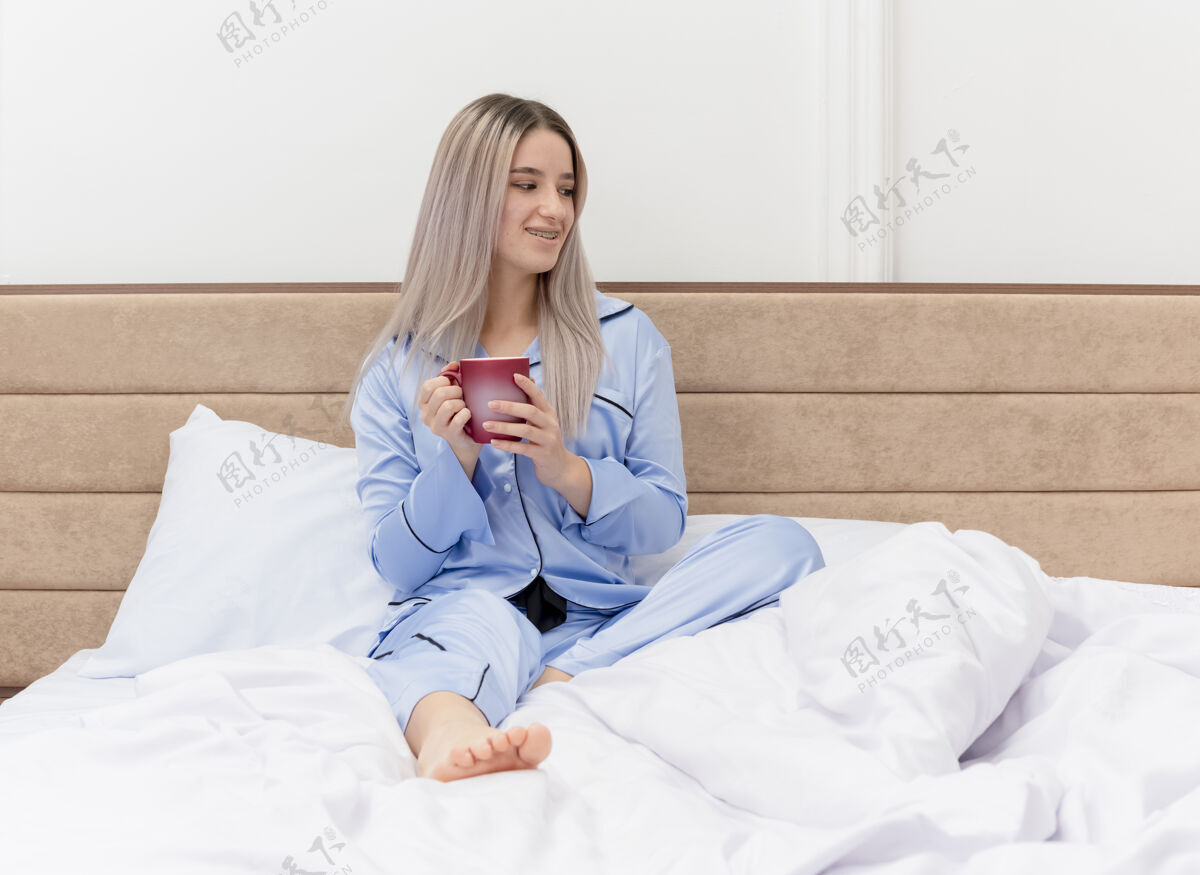 床穿着蓝色睡衣的年轻美女坐在床上 端着一杯咖啡 微笑着在卧室里享受早晨的时光时间年轻美丽