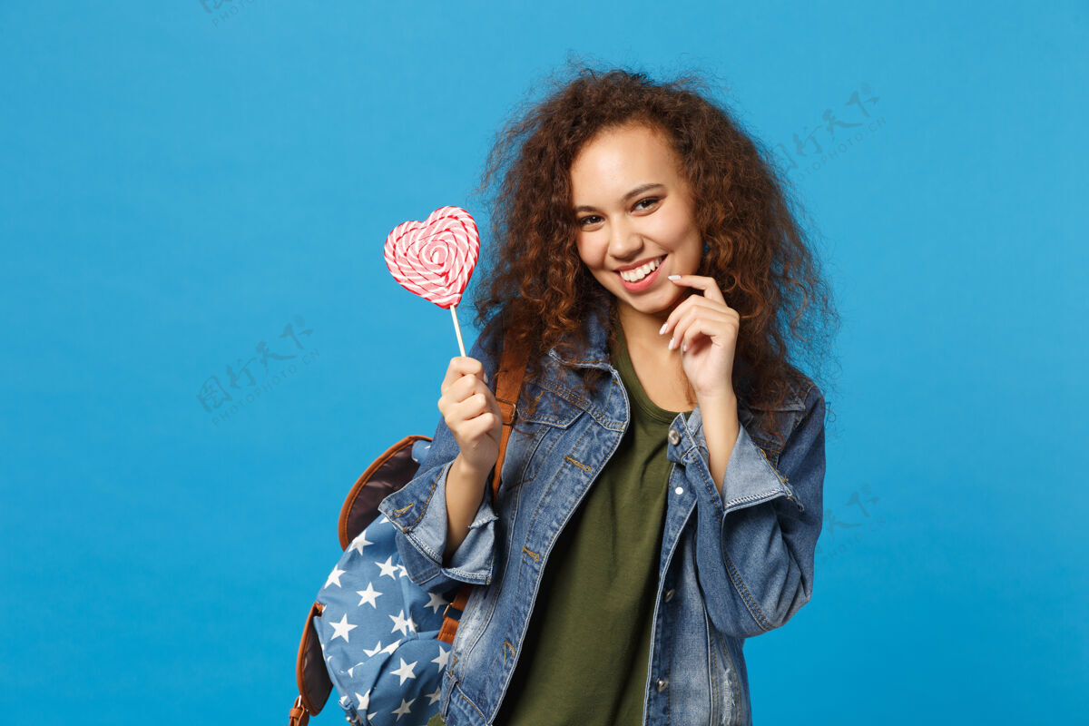 包年轻的非洲裔美国女孩十几岁的学生在牛仔服装 背包举行糖果隔离在蓝色的墙壁上好吃甜点糖