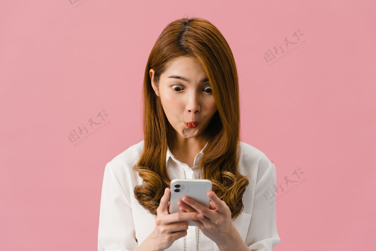 学生年轻的亚洲女士用手机 表情积极 笑容灿烂 穿着休闲服 感觉幸福 孤立地站在粉色背景上休闲媒体软件