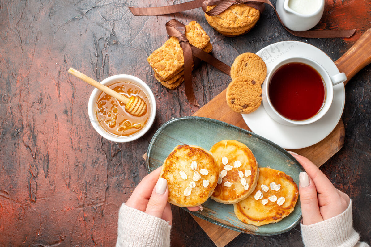 服用手拿托盘和新鲜煎饼的特写镜头一杯红茶放在木制砧板上蜂蜜饼干牛奶堆在黑暗的表面上牛奶蜂蜜晚餐