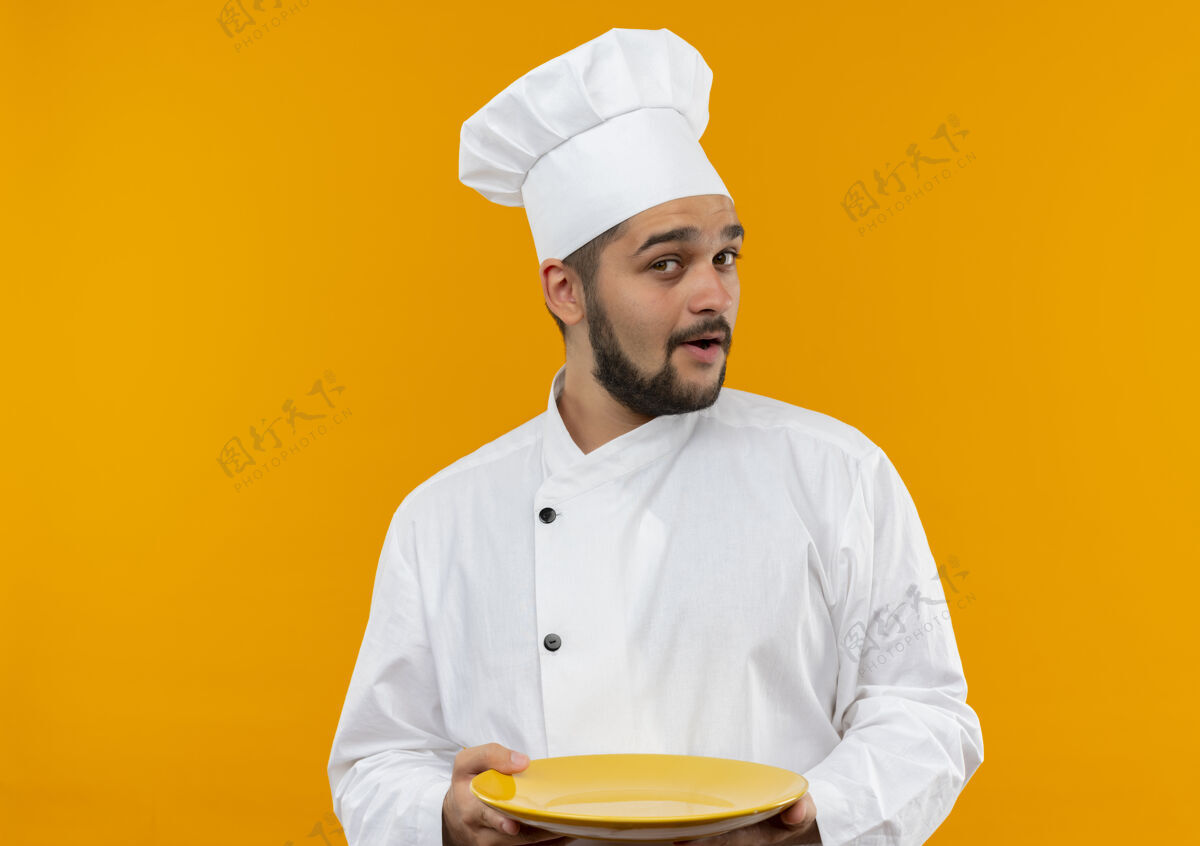 盘子令人印象深刻的年轻男厨师在厨师制服举行空盘子隔离在橙色墙上空制服烹饪