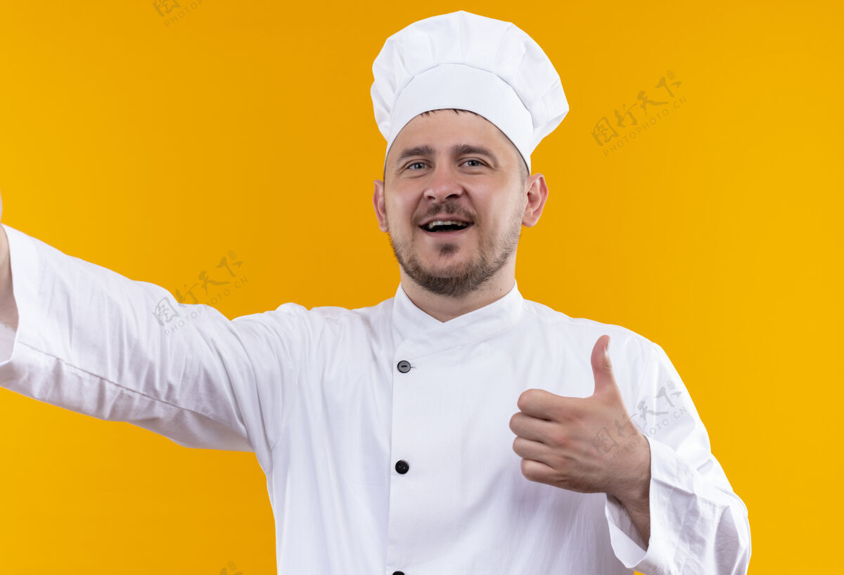 帅气身着厨师制服的年轻帅哥厨师在橙色的墙上孤立地竖起大拇指快乐年轻烹饪