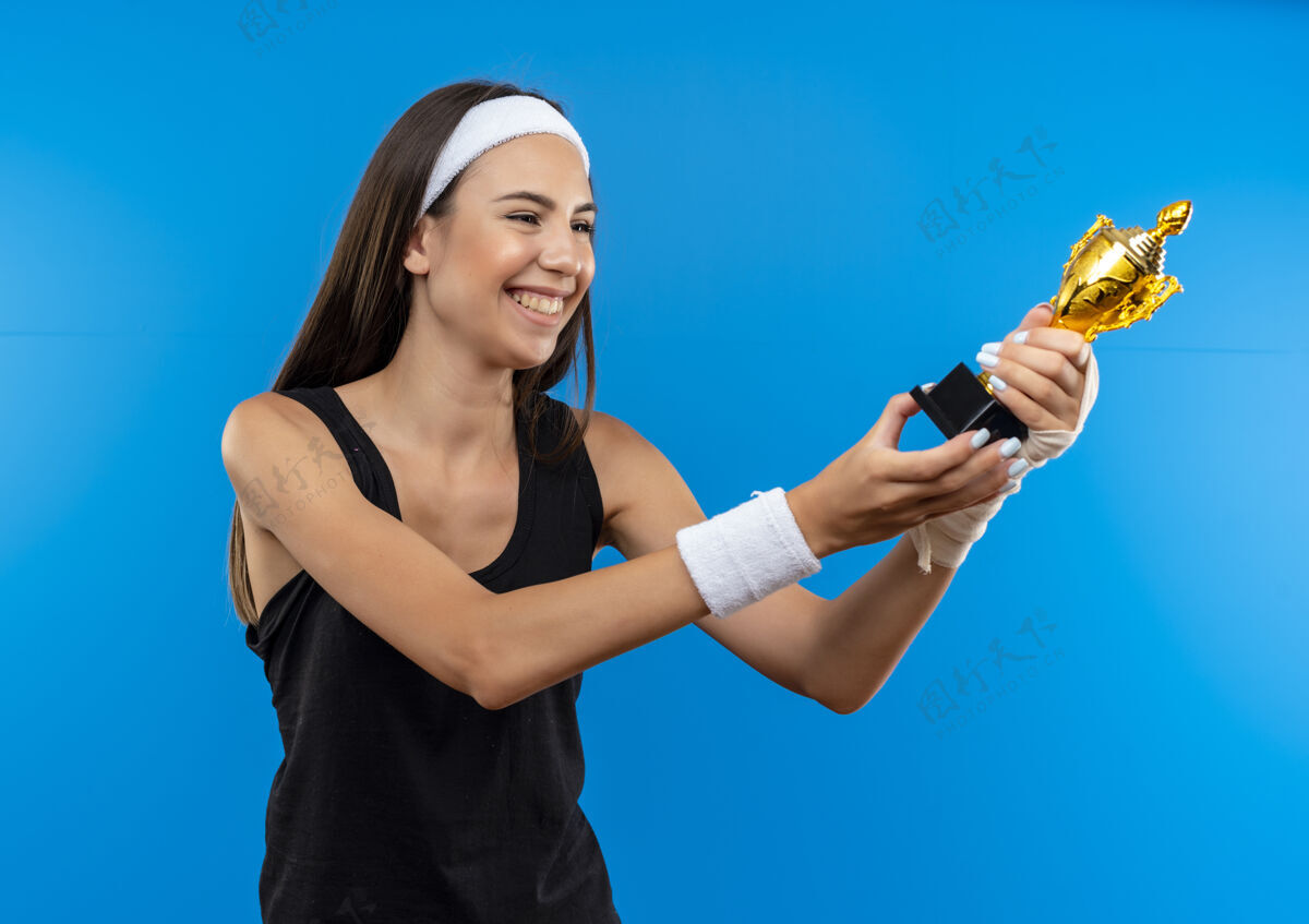 年轻快乐的年轻漂亮的运动女孩戴着头带和腕带拿着冠军杯看 一只手腕受伤 用绷带包着隔离在蓝色的墙上运动头带戴