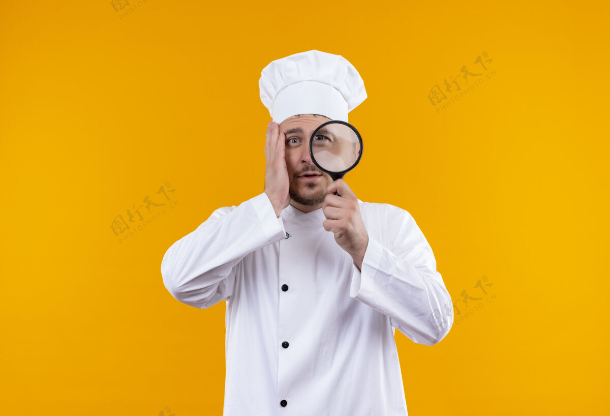 年轻年轻帅气的厨师穿着厨师制服 手放在放大镜上 脸贴在隔离的橙色墙上 留有复印空间 给人留下深刻印象玻璃手放大镜
