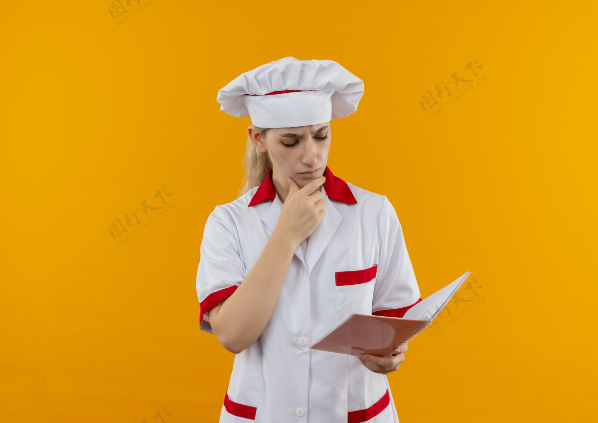 垫困惑的年轻漂亮的厨师穿着厨师制服 手放在下巴上 看着橘色墙上的便笺簿年轻厨师笔记