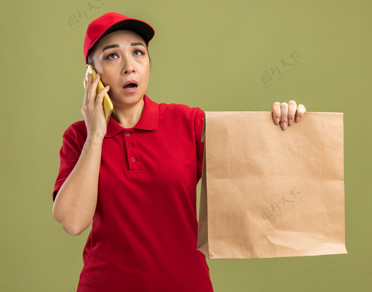 惊喜年轻的送货员身穿红色制服 戴着帽子 手里拿着一个纸包裹 站在绿色的墙上 一边用手机交谈 一边显得困惑和惊讶拿着帽子年轻