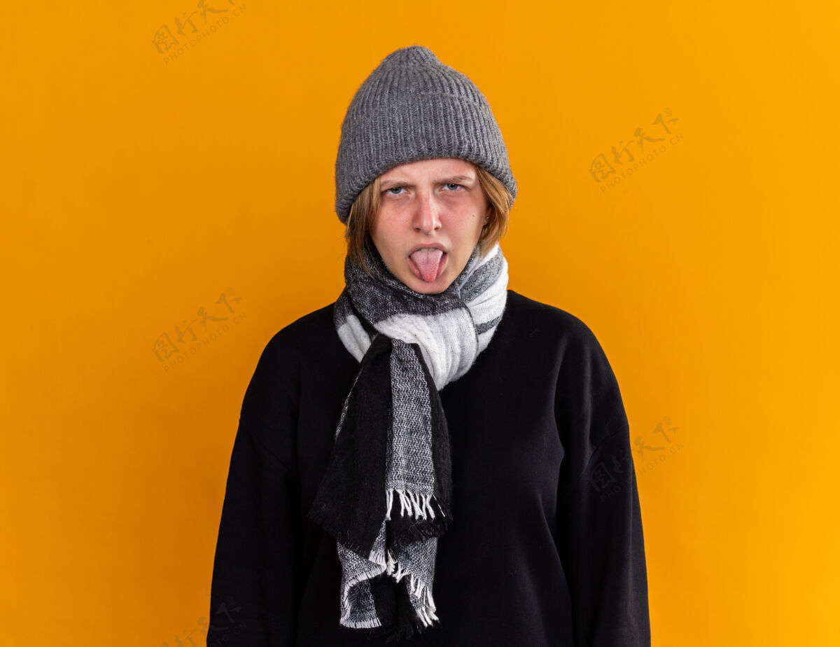 围巾不健康的年轻女子戴着暖和的帽子 脖子上围着围巾 感觉不舒服 患了感冒和流感 站在橙色的墙上吐出舌头 表情厌恶生病周围站