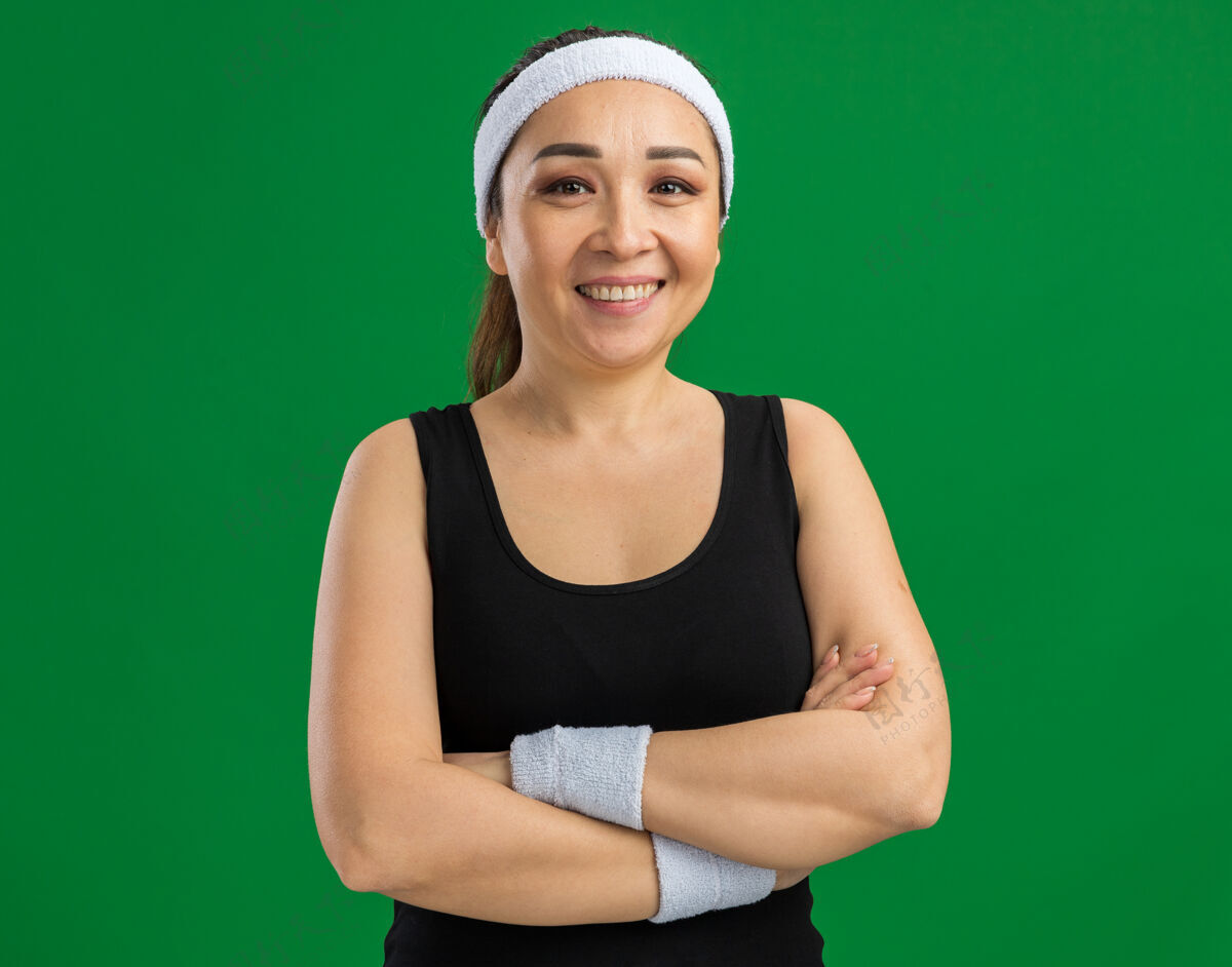 年轻戴着头巾和臂章的年轻健身女士 双臂交叉 自信地微笑着站在绿色的墙上臂章手臂健康