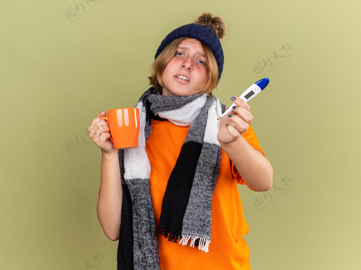 感冒不健康的年轻女子戴着帽子 脖子上围着围巾 感觉不舒服 喝着热茶 拿着数字体温计 患流感发烧 站在绿色的墙上发烧周围茶