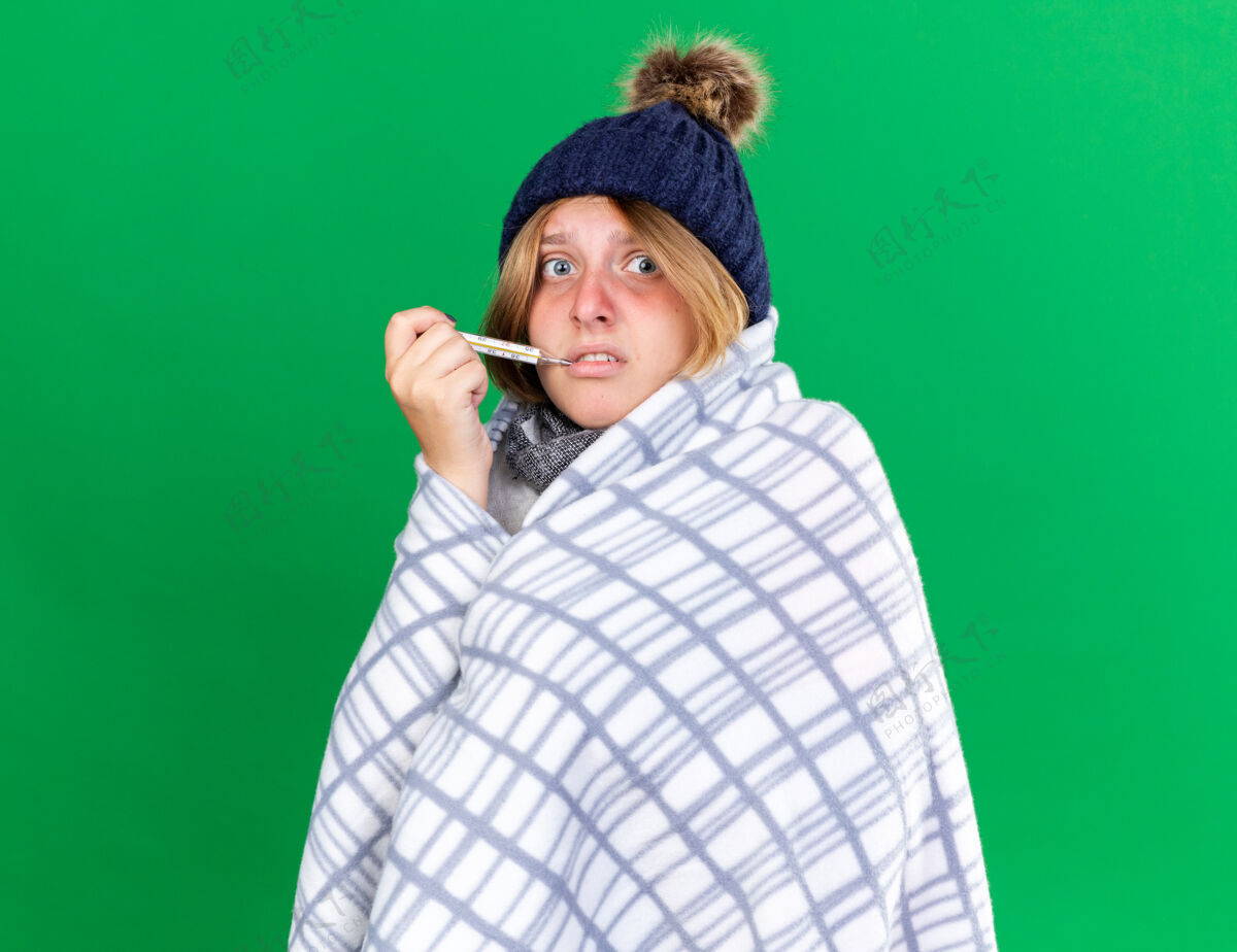 毯子不健康的年轻女子裹着毯子 戴着帽子 用体温计测量体温 患流感 发烧 站在绿色的墙上 看起来很担心体温计使用年轻人