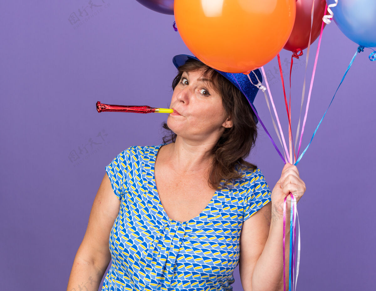 帽子快乐快乐的中年妇女戴着派对帽 手持一束五颜六色的气球吹着口哨 站在紫色的墙上庆祝生日派对吹束气球