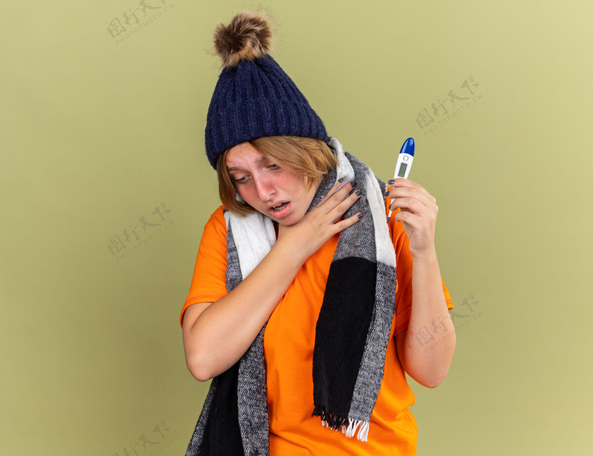 触摸不健康的年轻女子戴着帽子 脖子上围着围巾 感觉不舒服 拿着数字体温计 患流感 喉咙痛 站在绿色的墙壁上触摸她的脖子站感觉酸痛
