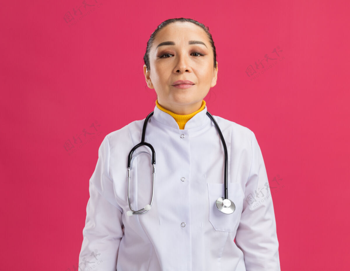 听诊器身穿白色药衣 脖子上戴着听诊器的年轻女医生站在粉红色的墙上 表情自信外套站表情