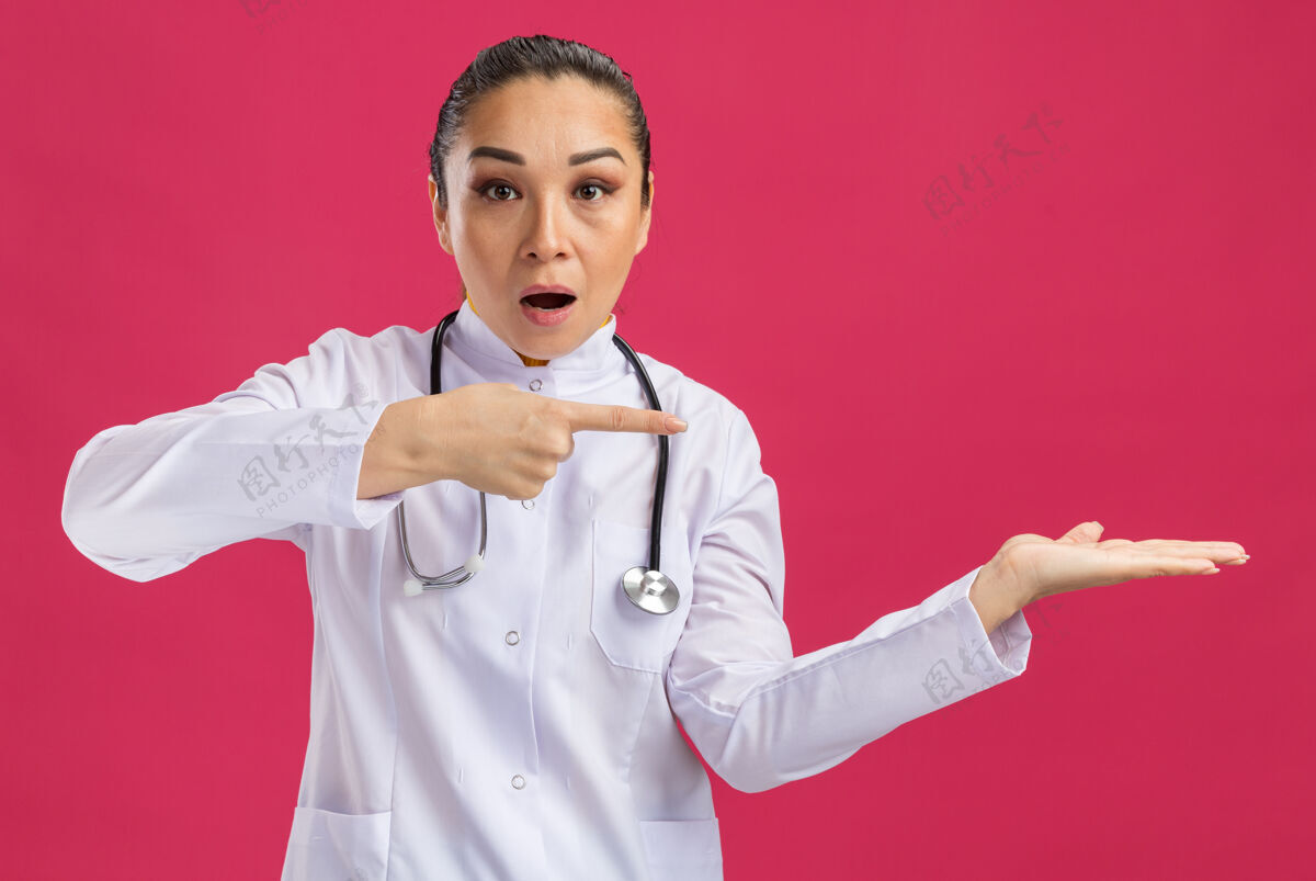 听诊器身穿白色药衣的年轻女医生 脖子上戴着听诊器 手臂指向复制区 食指站在粉红色的墙上站手臂周围
