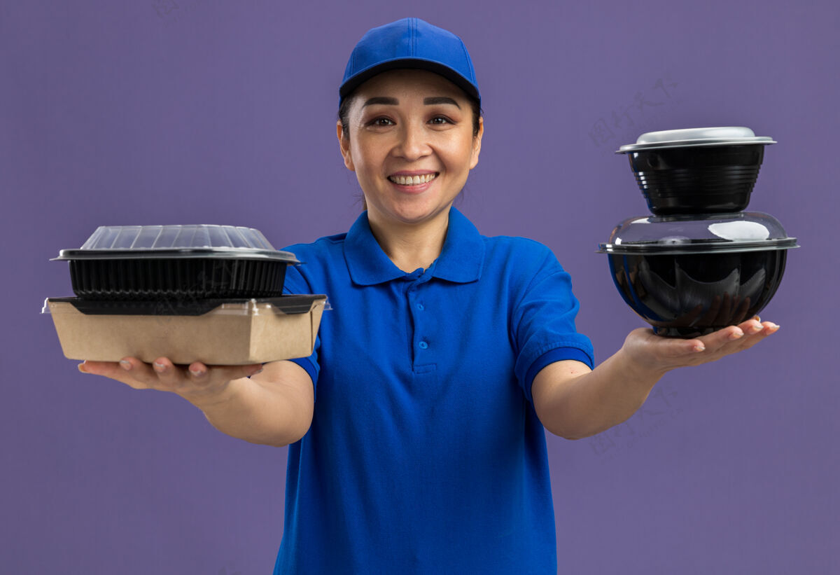 微笑身着蓝色制服 戴着帽子 手里拿着食品包 站在紫色的墙上 快乐地微笑着的年轻送货员食物年轻帽子