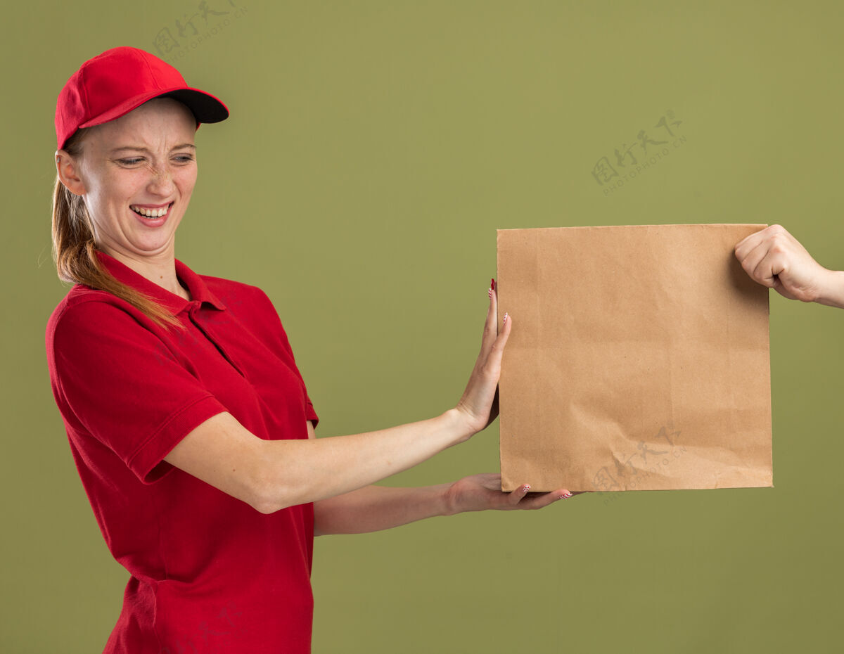 站立身穿红色制服 戴着帽子的年轻送货女孩站在绿色的墙上拒绝接受纸质包裹包装女孩帽子