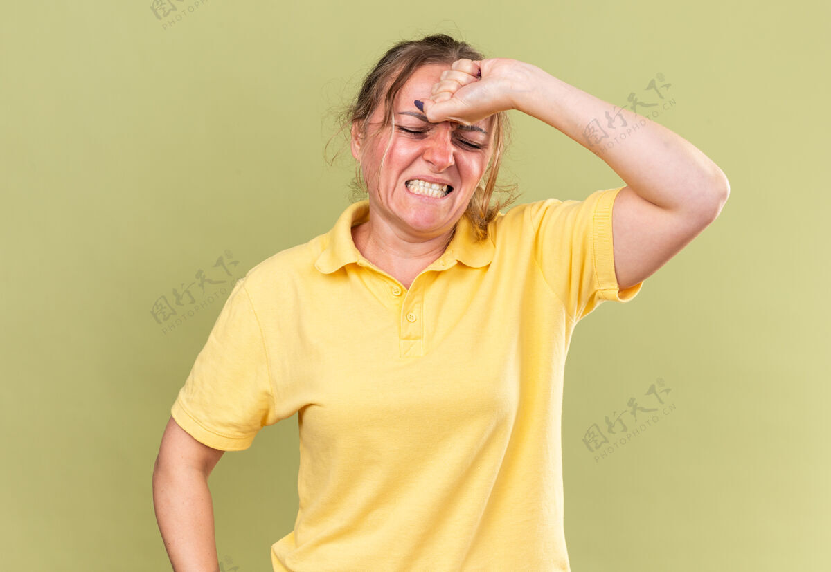 不健康一个穿着黄色衬衫的不健康的女人站在绿色的墙壁上摸着她的额头感到很难受 头痛得厉害 感冒可怕痛苦感情