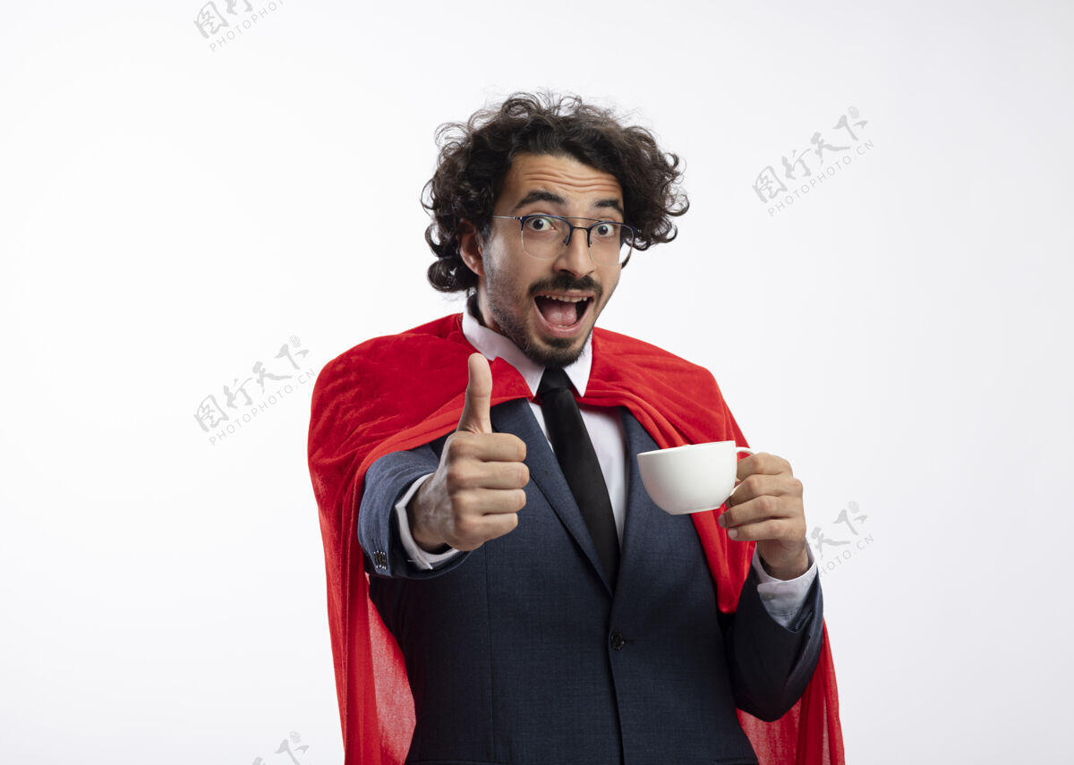 超级英雄惊讶的年轻白种人超级英雄戴着眼镜 穿着红色斗篷的西装 竖起大拇指 举着杯子红色光学穿