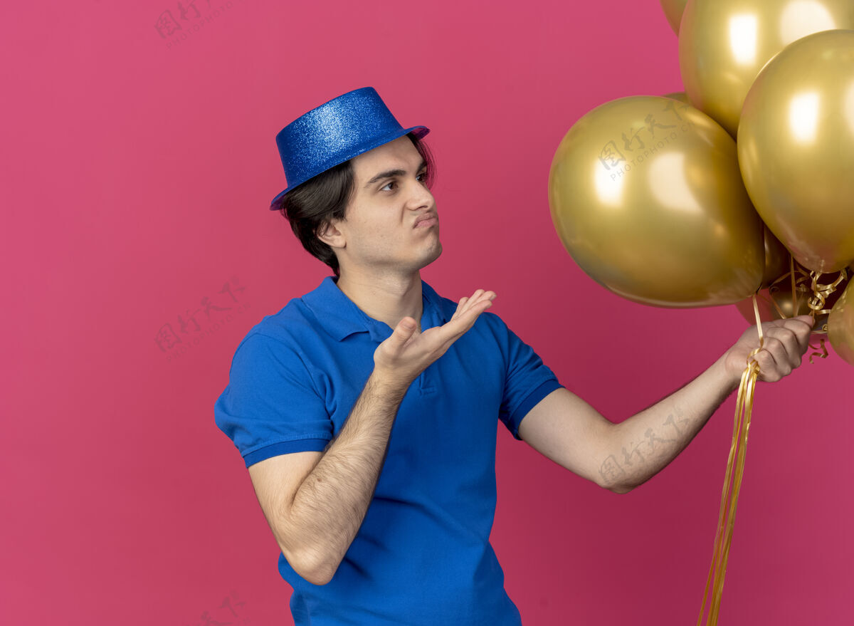 抱恼怒的白人帅哥戴着蓝色的派对帽指着氦气球空格蓝色粉色