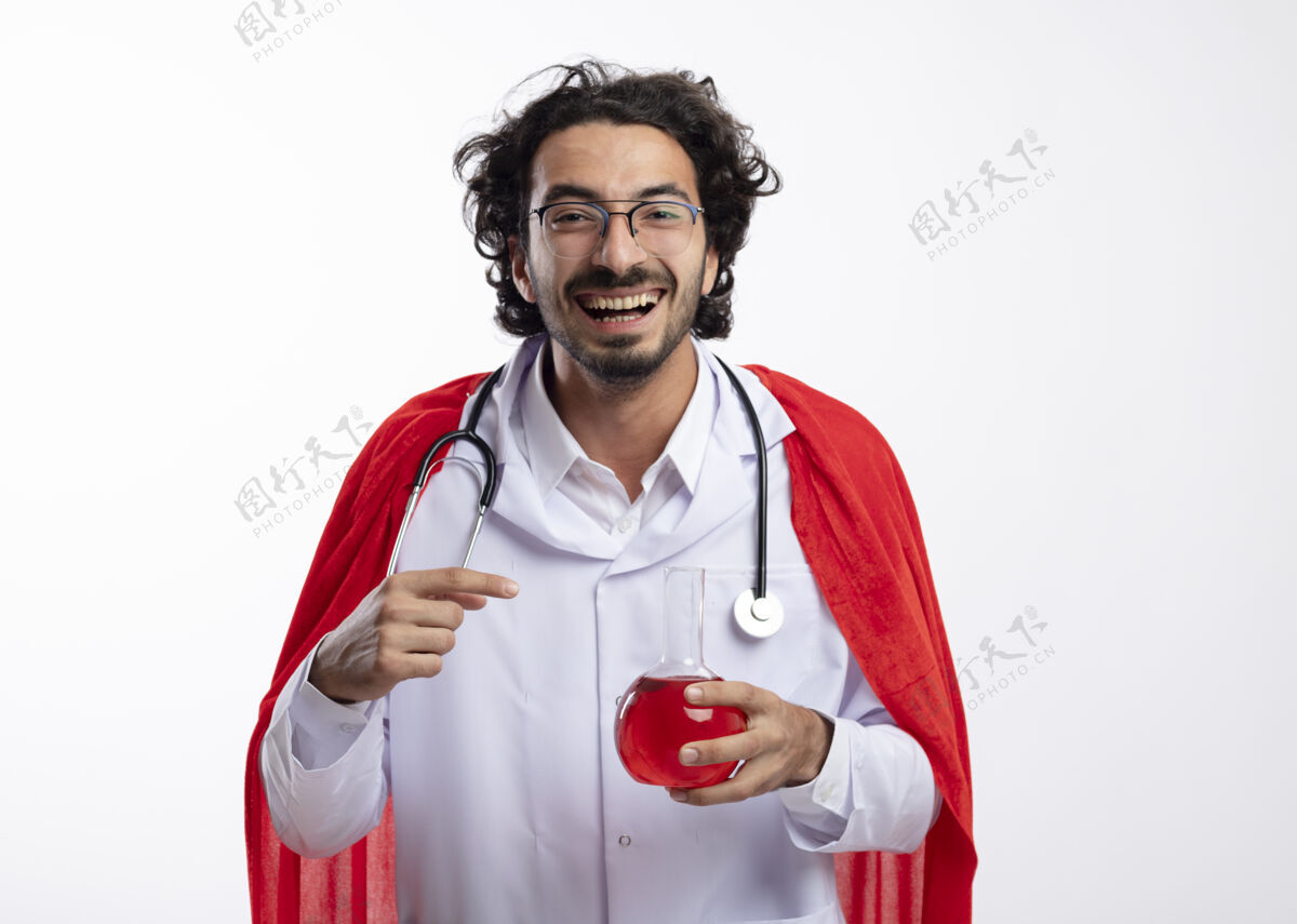 化学戴着眼镜 身穿医生制服 披着红色斗篷 脖子上戴着听诊器的快乐的白人年轻人握住并指着玻璃瓶中的红色化学液体烧瓶玻璃红
