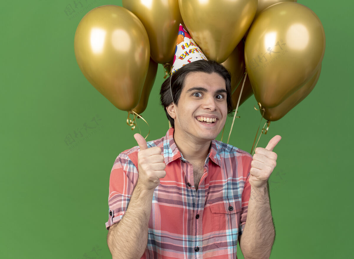 站着快乐英俊的白人男子戴着生日帽站在氦气球前双手竖起大拇指空间拇指复制