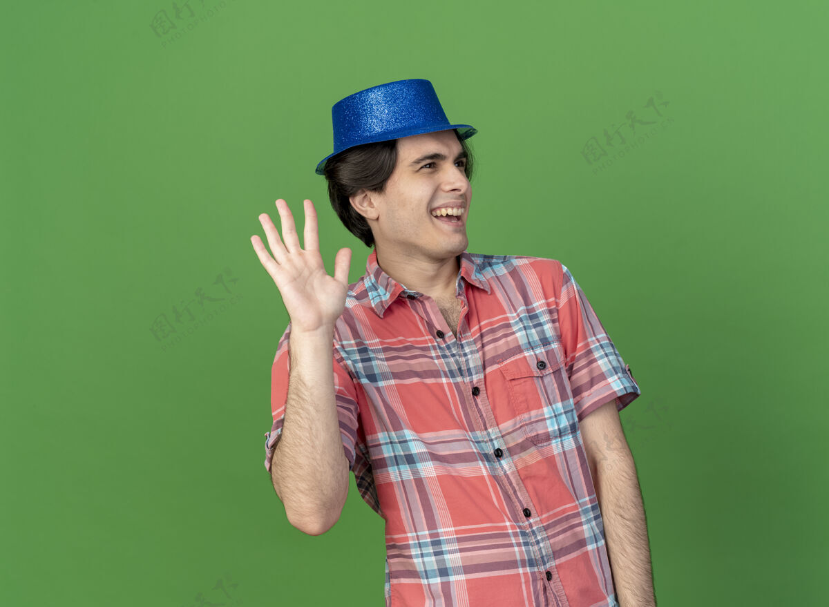 蓝色快乐英俊的白人男子戴着蓝色的派对帽站在那里 举起手看着一边男人提高穿