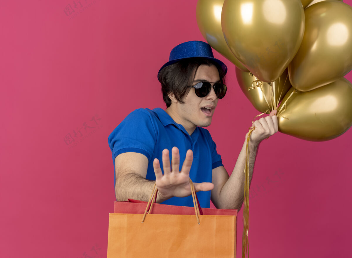 举行帅气的白种人戴着太阳眼镜 戴着蓝色的派对帽 手里拿着氦气球和纸购物袋 伸出手氦购物印象