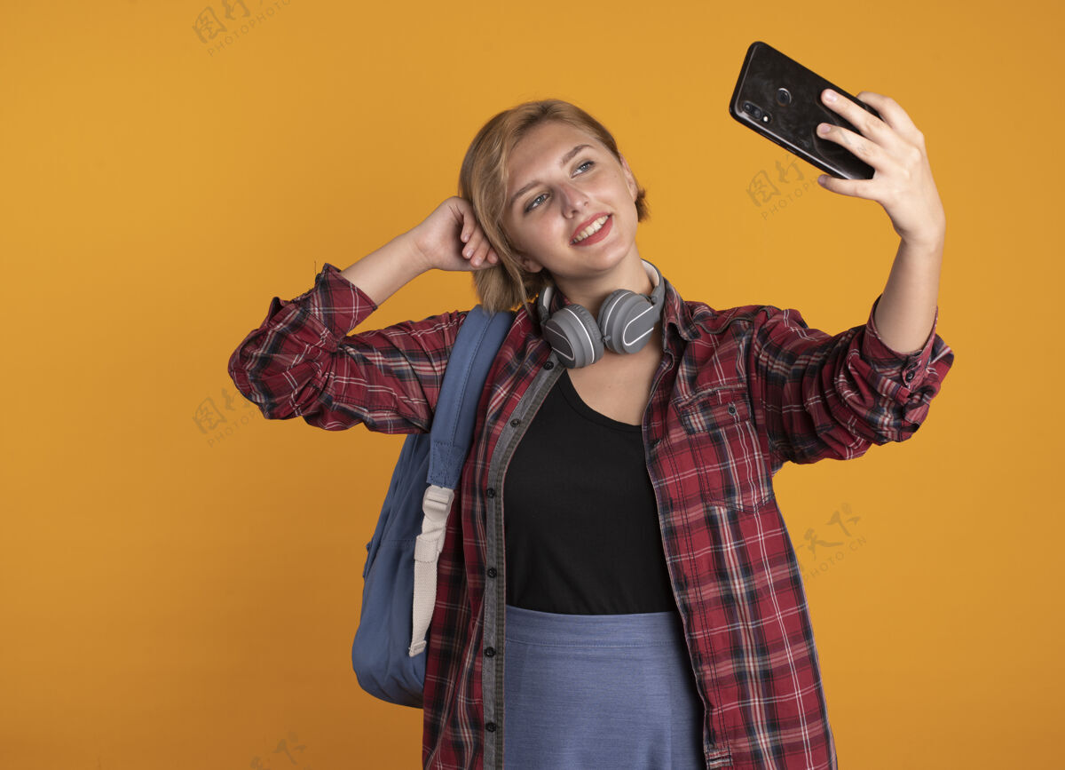 请高兴的年轻斯拉夫女学生戴着耳机 背着背包 手放在头上 拿着手机看斯拉夫背包橙色