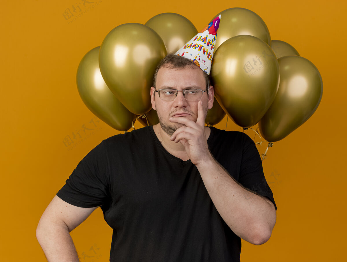 空间戴着生日帽 戴着光学眼镜的未被释放的成年斯拉夫男子把手放在脸上看着侧面 站在氦气球前壁板生日成人