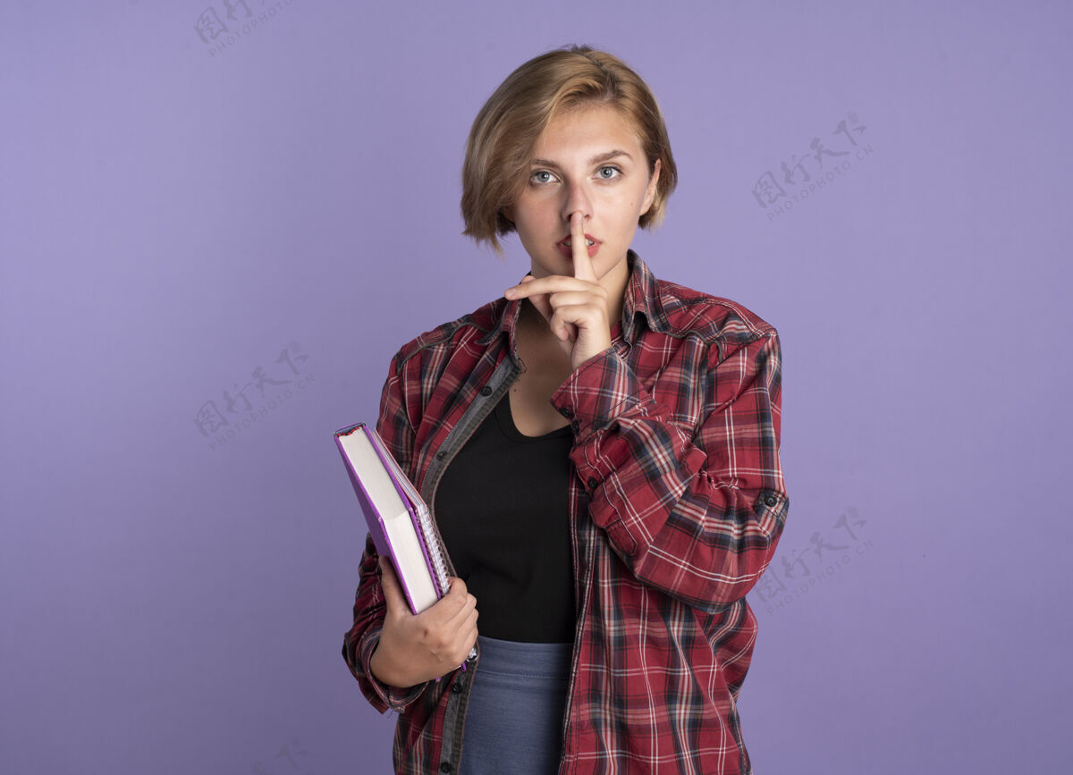 斯拉夫自信的年轻斯拉夫学生女孩拿着书和笔记本手势沉默的标志在紫色背景上与复制空间隔离笔记本紫色标志