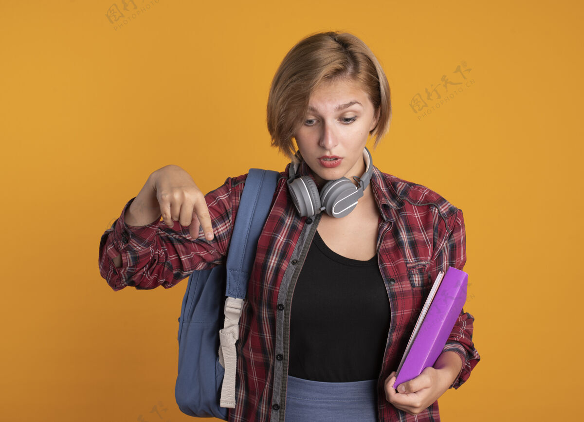 耳机惊讶的年轻斯拉夫女学生戴着耳机 背着背包 拿着书和笔记本朝下看复制惊喜橙色