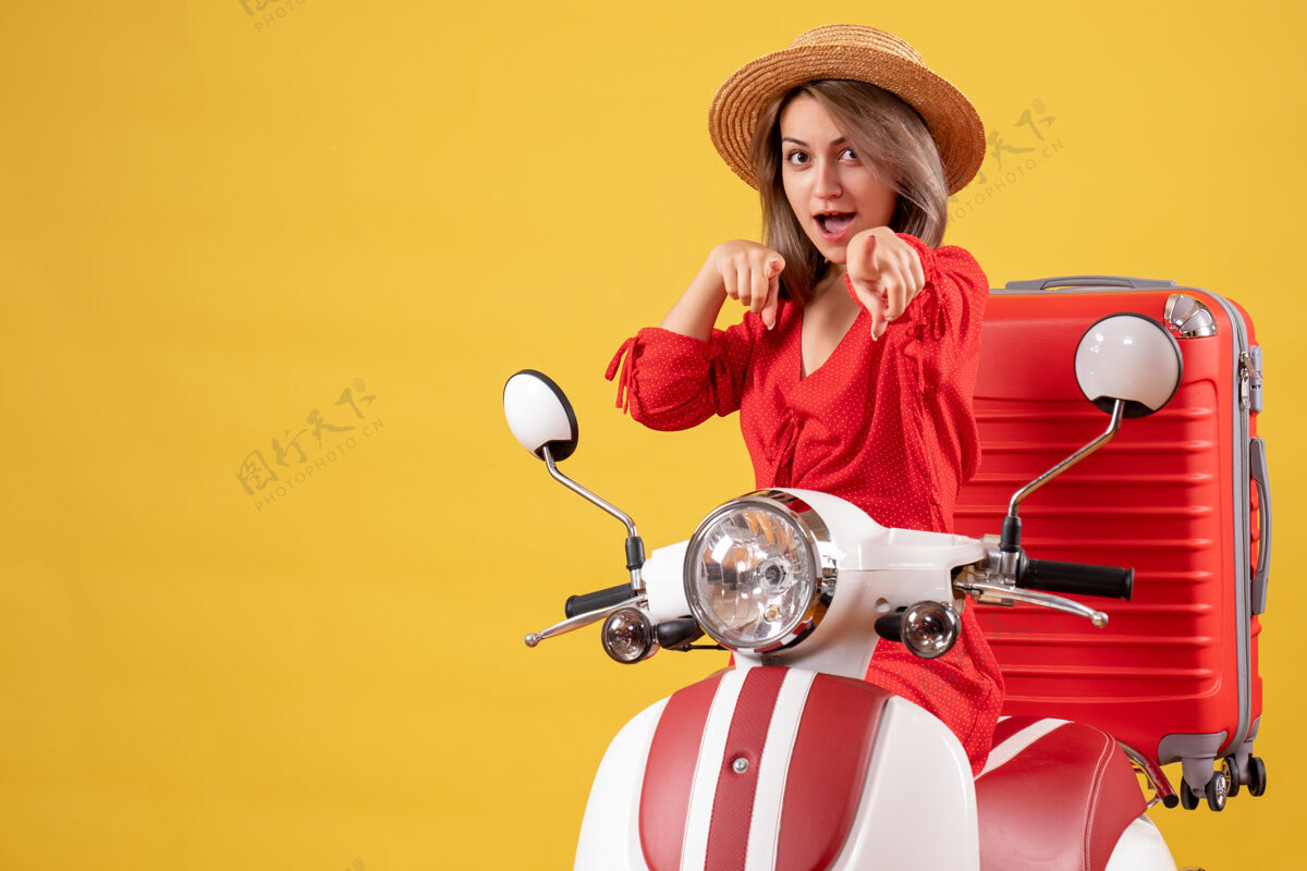 帽子骑着轻便摩托车的漂亮女孩 红色的手提箱指着摄像机车辆成人相机