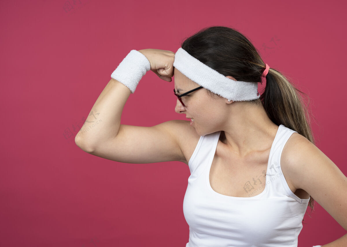 紧张愤怒的年轻运动女孩戴着眼镜戴着头带和腕带紧张的二头肌太空眼镜粉色