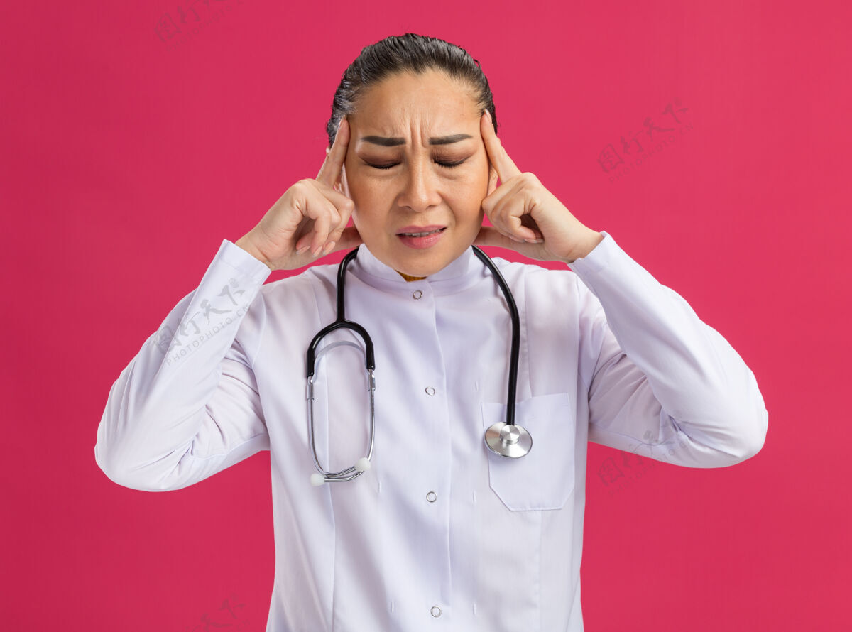 触摸身穿白色药衣 脖子上戴着听诊器的年轻女医生站在粉红色的墙上 用恼怒的表情抚摸着太阳穴 头痛头痛脖子站